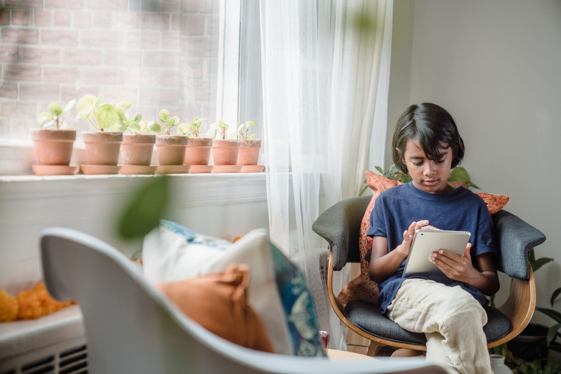 A child relaxing in a chair by a window at home, using a tablet